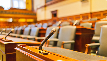 Close-up of a microphone on a wooden desk in an empty, wood-paneled parliament chamber or courtroom with rows of leather chairs in the blurred background