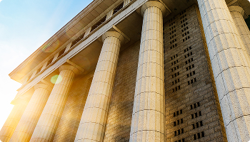 Low-angle view of a monumental building facade featuring massive classical stone columns illuminated by bright sunlight with a lens flare effect